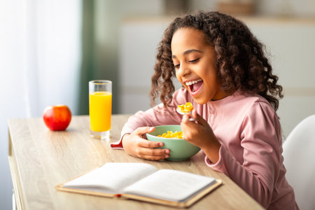 Cute black teen girl having healthy breakfast and reading book while sitting at desk at homeの写真素材