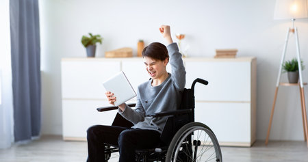 Excited teen boy in wheelchair holding tablet computer, celebrating his achievement or success,...