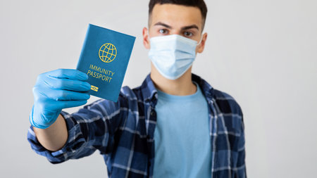 Young guy in medical mask showing covid-19 immunity passport on light studio background, selective focusの写真素材