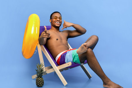 Flirty African American guy in swimwear showing CALL ME gesture, pointing at camera, sitting in lounge chairの写真素材