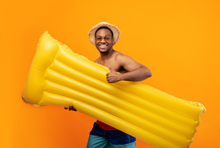Summertime beach fun. Positive black man in swimsuit going to pool with inflatable lilo, showing...