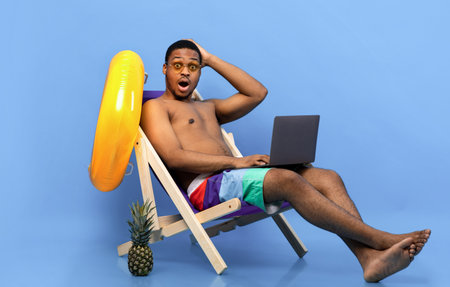 Shocked young black guy opening mouth and grabbing his head in surprise, using laptop in lounge chair, blue backgroundの写真素材