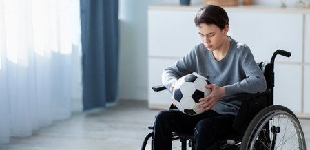 Full length of teen football player in wheelchair holding soccer ball and feeling stressed over his injury at homeの写真素材