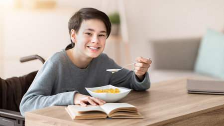 Positive impaired teen boy in wheelchair having breakfast and reading exciting book at home. Stay home activitiesの写真素材