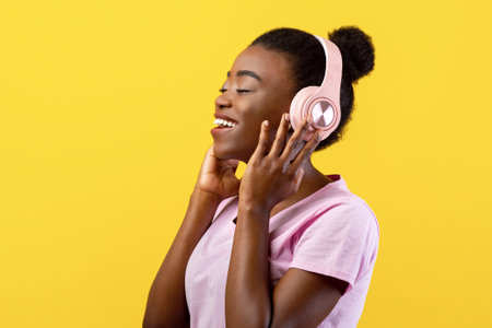 Favourite leisure. Carefree african american lady touching wireless headphones, listening to music on yellow backgroundの写真素材