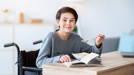 Cool disabled teenager in wheelchair having cereal for breakfast and reading book at home. Hobbies and pastimesの写真素材