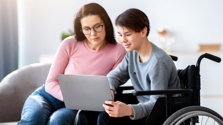 Remote education concept. Cheerful disabled teenager in wheelchair making online home assignment with his motherの写真素材