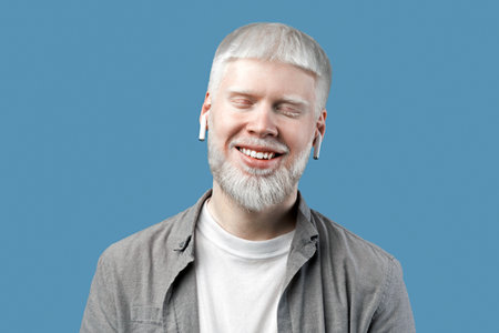 Relaxed albino guy listening music in bluetooth earpods, standing with closed eyes over turquoise backgroundの写真素材