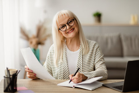 Smiling mature woman writing and holding document at homeの写真素材