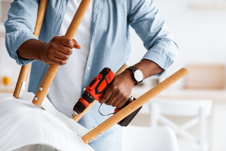 Furniture assembly process. African american handyman screwing legs to chair with electric screwdriver, cropの写真素材
