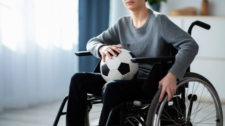 Cropped view of handicapped teen boy with soccer ball sitting in wheelchair, feeling upset over his injury at homeの写真素材