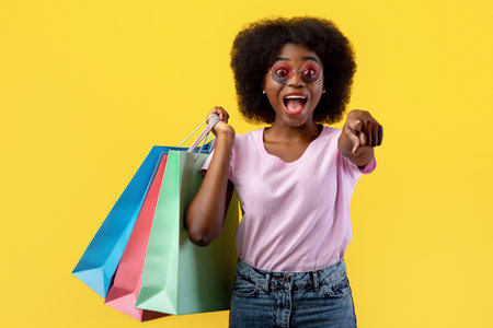 Hey, you. Emotional black woman pointing at camera and holding colorful shopping bags standing over yellow backgroundの写真素材