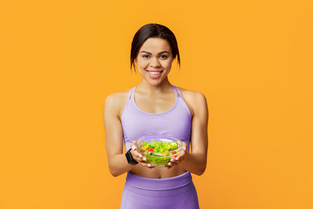Excited african american fit woman holding bowl with fresh vegetable salad standing on yellow studio...