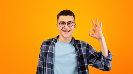 Portrait of happy millennial guy in glasses showing OK gesture over orange studio backgroundの写真素材