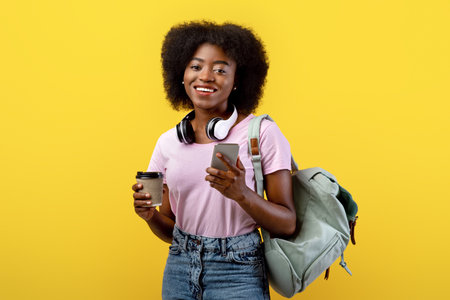 Positive african american woman with backpack and smartphone holding coffee to go over yellow backgroundの写真素材