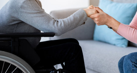 Handicapped teenage boy in wheelchair receiving support from his mother or caregiver, holding her hands at home, closeupの写真素材