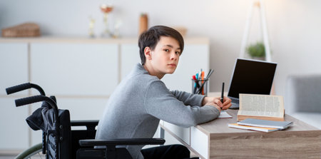 Disabled teen boy in wheelchair studying online with textbooks and laptop at home, mockup for design on pc screenの写真素材