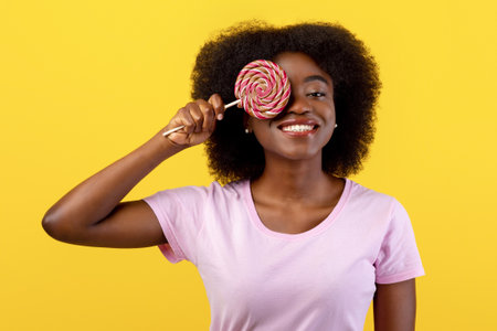 Sweet tooth. Positive young african american woman covering one eye with lollipop over bright yellow studio backgroundの写真素材