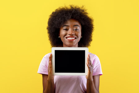 African american lady demonstrating digital tablet with free screen, mockup for text or advertisement, yellow backgroundの写真素材
