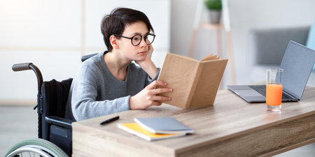 Bored teen boy in wheelchair reading textbook, making dull home assignment near laptop indoorsの写真素材