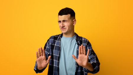 Disgusted millennial guy showing STOP gesture with both hands, expressing rejection on orange studio...