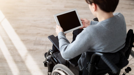 Handicapped teenage boy in wheelchair holding tablet with blank screen indoors, mockup for website or app designの写真素材