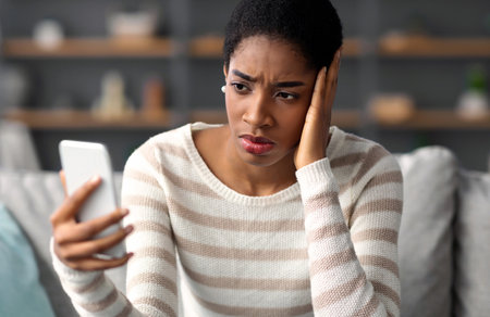 Worried Black Woman Looking At Smartphone While Sitting On Couch At Homeの写真素材