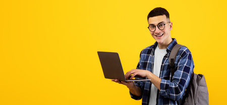Intelligent young student with backpack using laptop pc on orange studio background