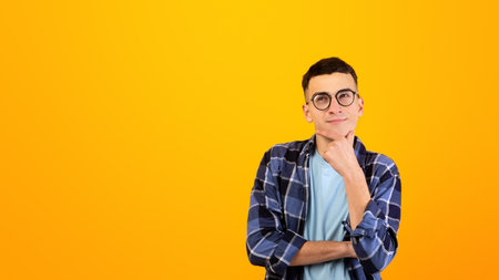 Pensive young man in glasses touching his chin, thinking over something on orange studio background