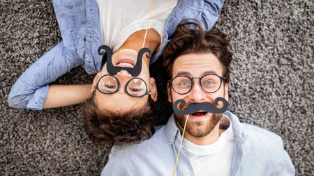 Joyful father and his son having fun with fake moustache on sticks, wearing glasses and smiling to...