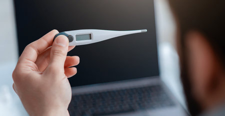 Sick during work from home. Man holding digital thermometer in front of laptop, selective focus.