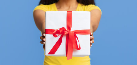 Unrecognizable joyful black woman stretching wrapped gift box to camera, standing over blue studio...