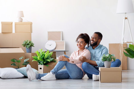 Cheerful young black husband and wife rest, sit on floor among cardboard boxes with things and...
