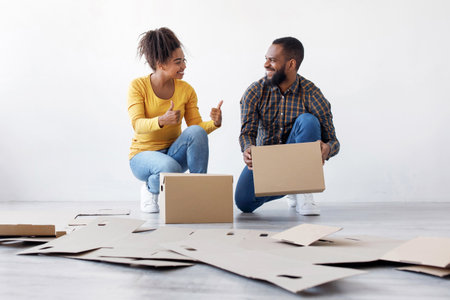 Smiling young black couple stack cardboard boxes for moving, lady show thumbs up sign in empty room