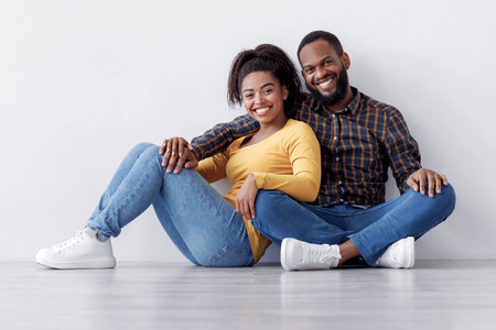 Happy millennial african american couple hugging sits on floor in empty room on white wall...