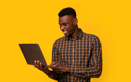 Freelance Work. Portrait Of Smiling African Guy Using Laptop Over Yellow Background