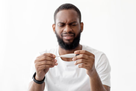 Unhappy sad young black bearded man hold thermometer with high temperature in bedroom