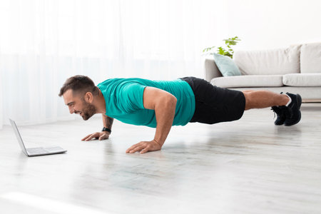Cheerful millennial muscular european man doing push ups and watching online lesson on computer in...