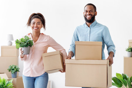 Satisfied young black family carry cardboard boxes and potted plants in white room interior, free...