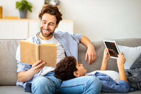 Different generations. Father reading book while his son using digital tablet, sitting together on...