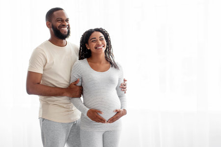 Cheerful satisfied handsome young black man hugs pregnant woman with belly in bedroom interior