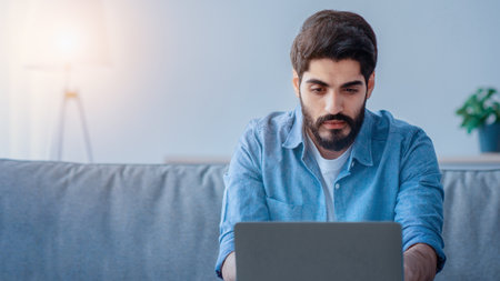 Modern job. Concentrated arab man working on laptop at home office, sitting on sofa and using...