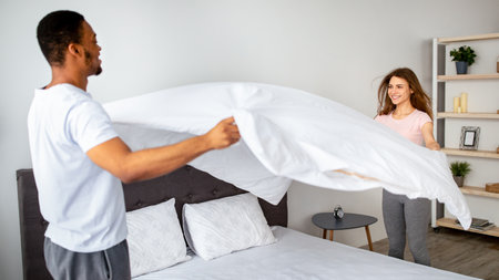 Domestic chores. Millennial multinational couple in love making bed at home, having fun while doing housework togetherの写真素材