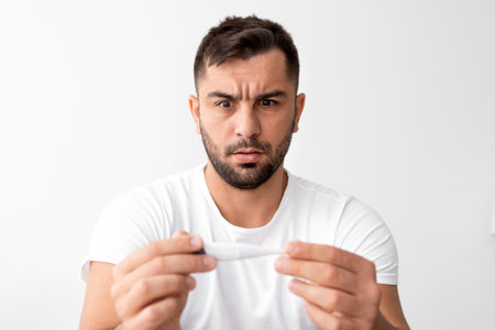 Sad shocked young male with beard looks at thermometer with high temperature and suffering from ill...