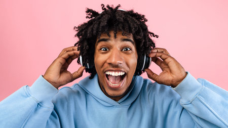 Overjoyed black teenager enjoying popular music in headset, opening mouth in excitement on pink...