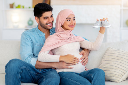Loving arab man hugging his pregnant wife with baby booties, sitting together on sofa at home