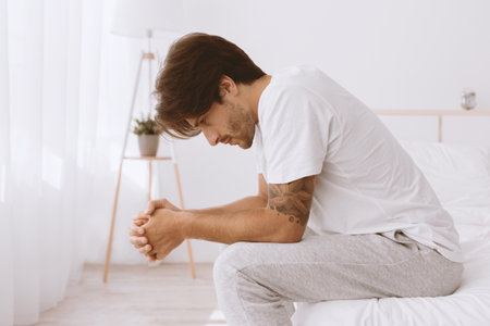 Upset caucasian young husband sitting in bed, suffering from weakness, wife ignoring him in white bedroomの写真素材