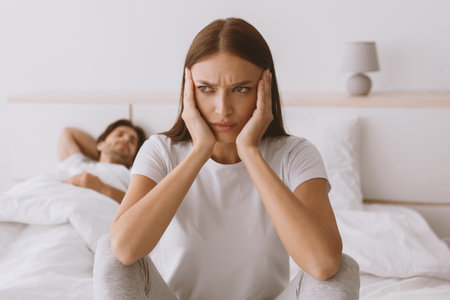 Offended sad upset pretty young caucasian woman sits on bed while husband sleeps in bedroom interiorの写真素材
