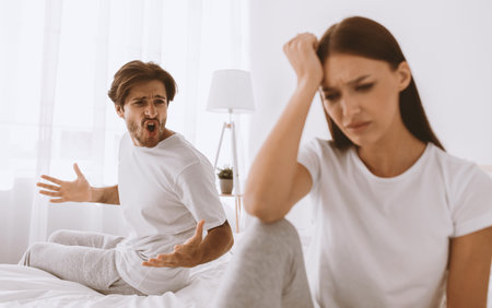 Aggressive evil millennial caucasian husband with stubble shouts and swears at offended sad depressed wife in bedroomの写真素材