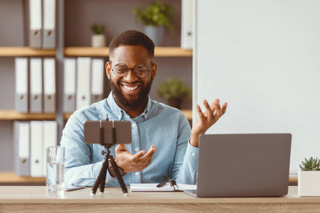 Smiling millennial black male teacher in glasses look at laptop, phone webcam in home office...